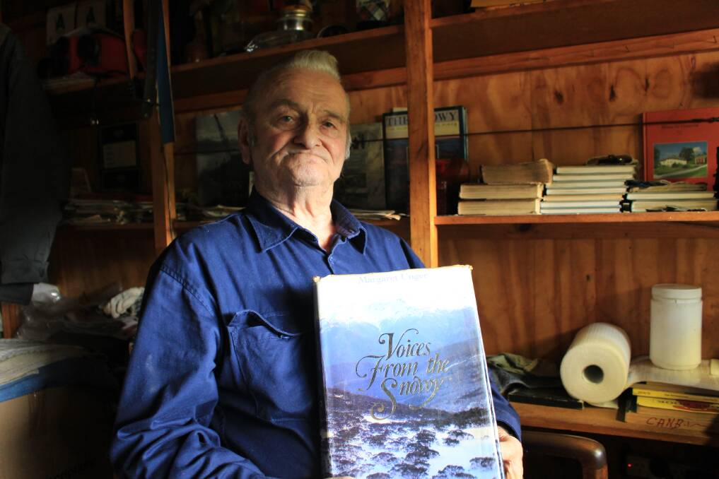 82-year-old Reidar Herfoss in his workshop in Eden with a book about the Snowy Hydro Scheme that features a photo of Reidar back in his glory days. 
